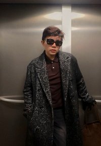 Szary płaszcz w jodełkę z dużymi klapami; ciemne okulary przeciwsłoneczne; brązowa koszulka polo; trzyma brązową torbę; stoi w windzie.