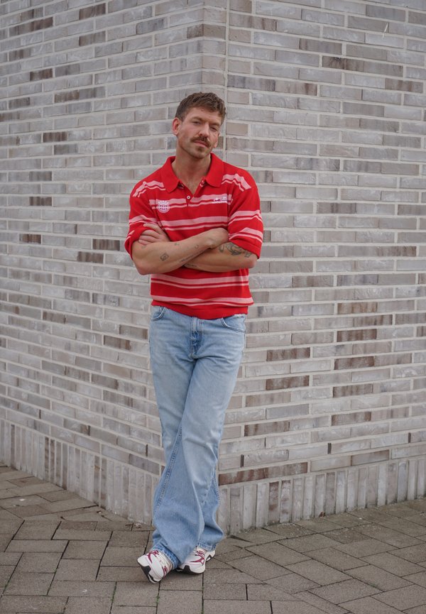 Homme aux cheveux courts et tatouages portant un polo rayé rouge, un jean bleu clair et des baskets, appuyé contre un mur en briques grises, les bras croisés.