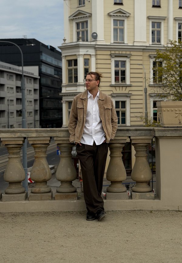 Hombre con chaqueta beige y camisa blanca, de pie junto a una barandilla de piedra con edificios históricos y una calle de la ciudad al fondo.