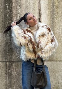 Femme avec une longue queue de cheval noire portant une veste en fourrure crème et marron, un col roulé beige, un jean bleu, tenant un sac à main tressé foncé devant un mur texturé.