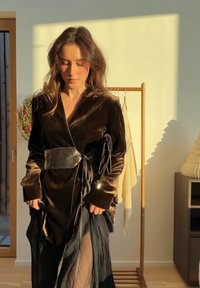 Femme portant une robe portefeuille en velours marron et une jupe noire transparente, debout à l'intérieur près d'un porte-vêtements en bois avec un vêtement beige.