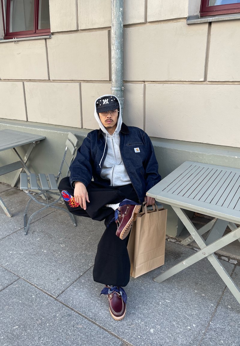 Homme portant une casquette, des lunettes, une veste en jean et des bottes est assis sur le trottoir entre deux tables d'extérieur grises, tenant un sac en papier brun.