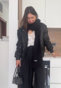 Femme debout dans une cuisine, portant une veste noire en cuir, une écharpe noire, un haut en dentelle blanc, un pantalon noir, tenant un sac à main noir.