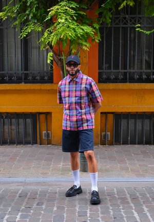 Camisa de cuadros de manga corta en azul, rosa y blanco, combinada con pantalones cortos de mezclilla oscura. Zapatos negros, calcetines blancos y una gorra negra. Entorno urbano.