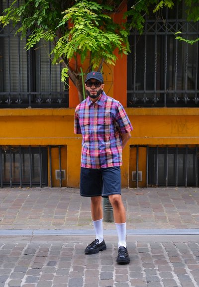 Camisa de cuadros de manga corta en azul, rosa y blanco, combinada con pantalones cortos de mezclilla oscura. Zapatos negros, calcetines blancos y una gorra negra. Entorno urbano.