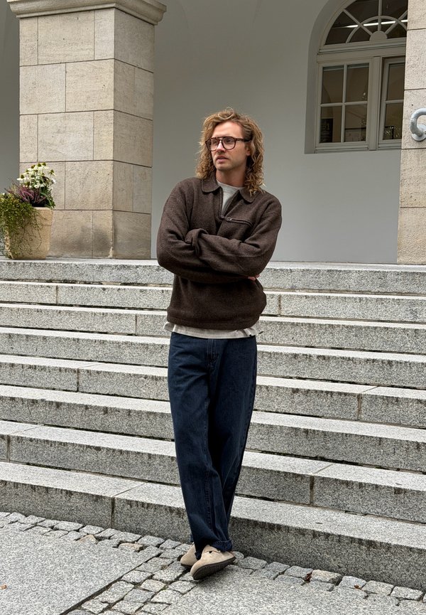 Hombre con gafas y cabello rizado de pie con los brazos cruzados junto a unos escalones de piedra cerca de una maceta con flores y un fondo de ventana arqueada.
