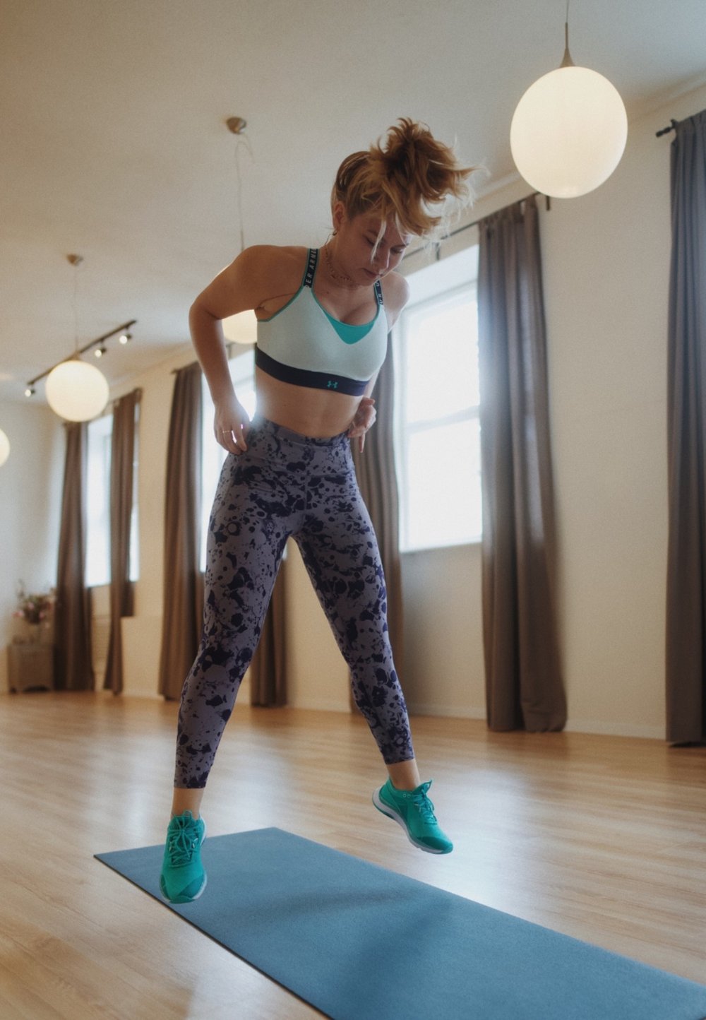 Sport-BH in hellblau und schwarz, gemusterte lila Leggings, türkisfarbene Sneakers, auf einer blauen Matte in einem großzügigen Innenraum mit Holzboden.