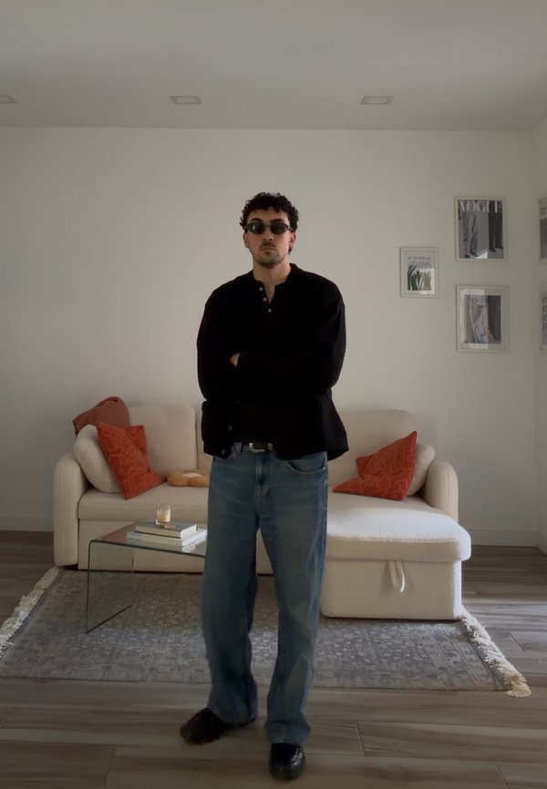 Uomo con occhiali da sole, maglietta nera e jeans, in piedi con le braccia incrociate in un soggiorno con un divano bianco e cuscini arancioni.