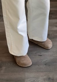 Tan suède slippers met ronde neuzen, gestikte details en een zachte textuur, gedragen met witte wijde broek op een houten vloer.