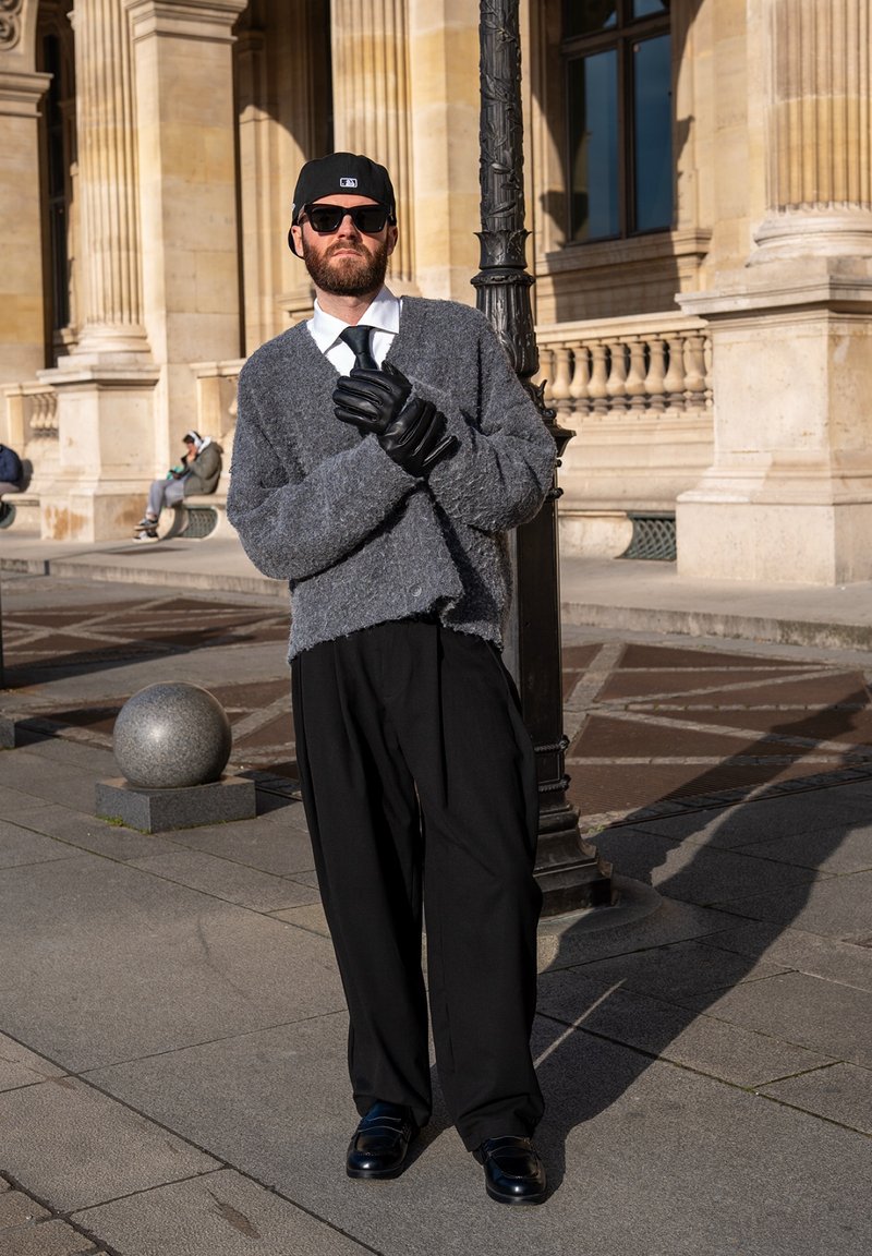 Homme portant des lunettes de soleil, une casquette noire, un cardigan gris, des gants noirs et un pantalon habillé, se tenant dans une rue de la ville près d'un bâtiment en pierre historique sous la lumière du soleil.