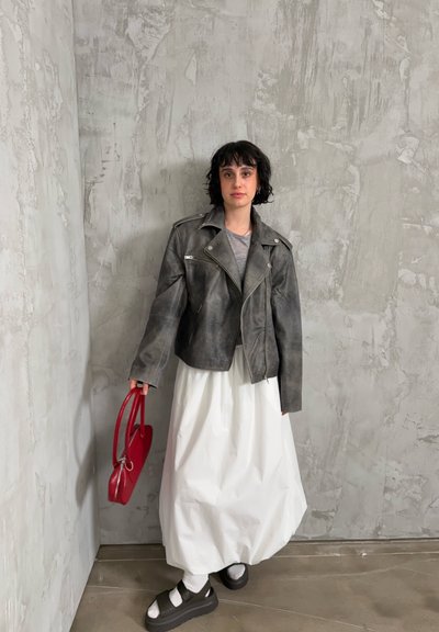 Persona con chaqueta de cuero gris, falda larga blanca, sandalias negras con plataforma y calcetines blancos, sosteniendo bolso rojo, de pie contra una pared gris texturizada.