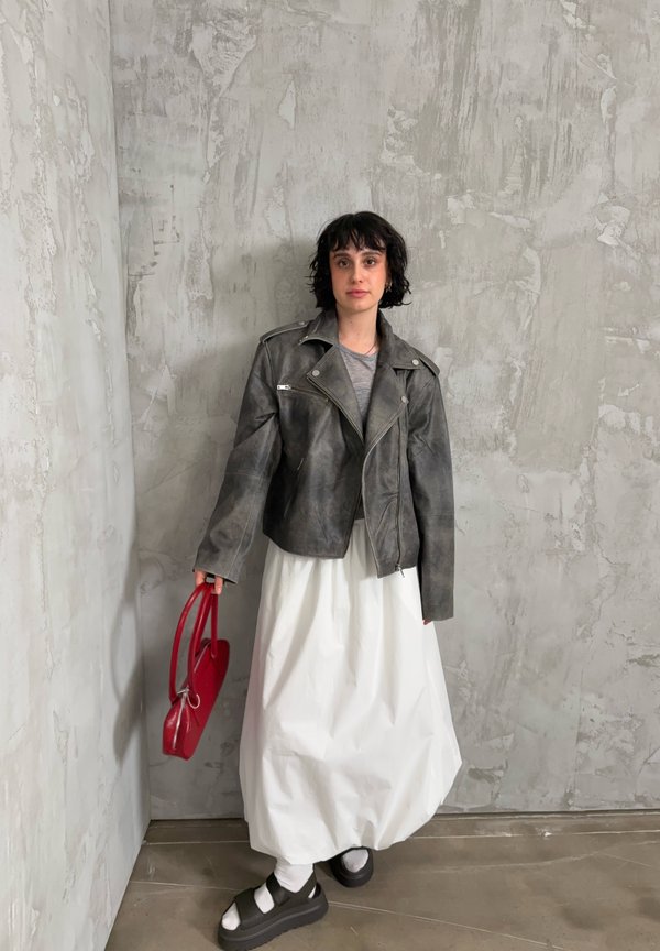 Osoba oblečená v šedej koženej bunde, bielej dlhej sukni, čiernych platformových sandáloch s bielymi ponožkami, držiaca červenú kabelku, stojaca pred textúrovanou sivou stenou.