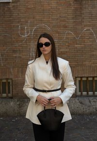 Blazer alb croitorit cu guler înalt, talie neagră cu curea, asortat cu ochelari de soare rotunzi negri și o geantă mică neagră, purtat cu pantaloni negri.