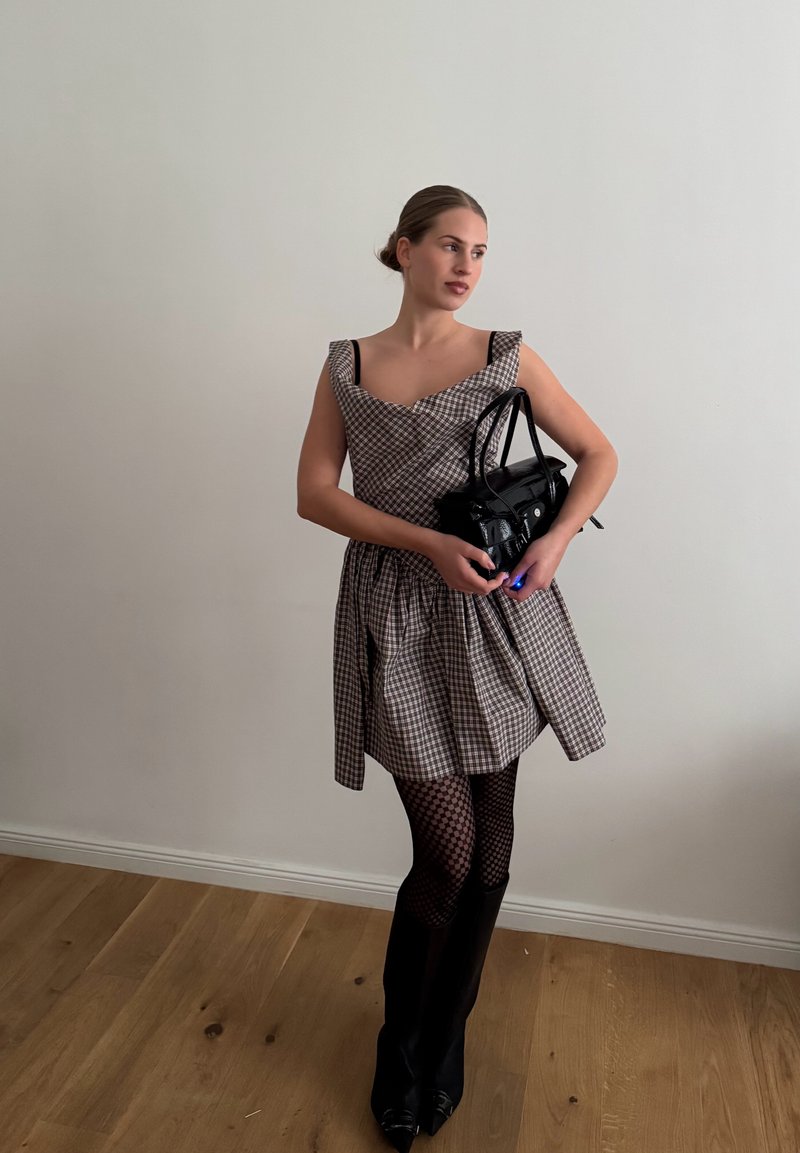 Femme en robe à carreaux, collants à motif et bottes noires tenant un sac à main noir, debout sur un sol en bois contre un mur blanc uni.