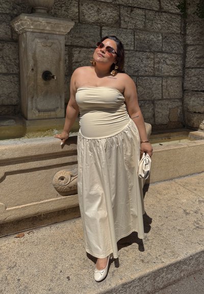 Vestido crema sin tirantes con un corpiño ajustado y falda fluida, combinado con bailarinas blancas y un clutch a juego. Entorno al aire libre iluminado por el sol.