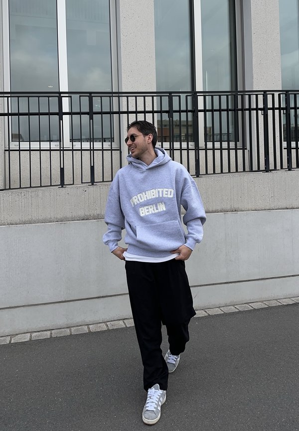 Άνδρας που περπατάει στο πεζοδρόμιο φορώντας γυαλιά ηλίου, μια γκρι μπλούζα με κουκούλα "PROHIBITED BERLIN", μαύρο παντελόνι και γκρι αθλητικά παπούτσια κοντά σε ένα μοντέρνο κτίριο.