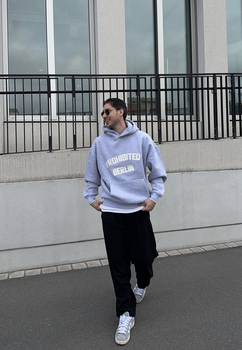 Vīrietis pastaigājas trotuārī, valkājot saulesbrilles, pelēku kapuci ar uzrakstu "PROHIBITED BERLIN", melnas bikses un pelēkus sporta apavus netālu no modernas ēkas.