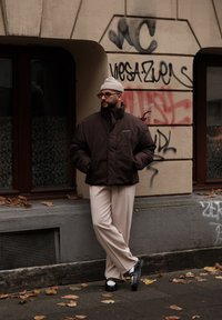 Καφέ φουσκωτό μπουφάν, κρεμ παντελόνι, μαύρες πλατφόρμες. Στηρίζει τον εαυτό του σε έναν τοίχο με γκράφιτι, με φθινοπωρινά φύλλα στο έδαφος.