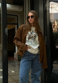 Casaco de camurça castanho sobre uma t-shirt branca com um gráfico e texto azul, combinado com uns jeans azuis largos, accessorized com óculos de sol escuros.