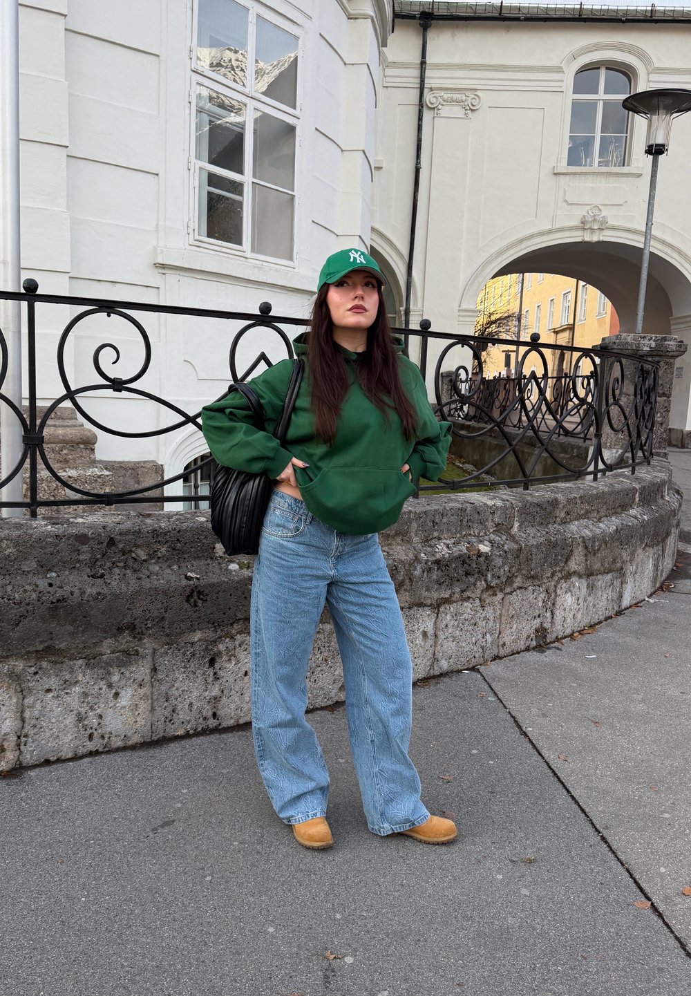 Sweat-shirt à capuche vert, jeans larges bleu, sabots beige, sac à main noir. Casquette verte avec logo. Mur en pierre et architecture en arrière-plan.