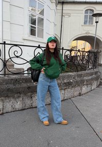Sweat-shirt à capuche vert, jeans larges bleu, sabots beige, sac à main noir. Casquette verte avec logo. Mur en pierre et architecture en arrière-plan.