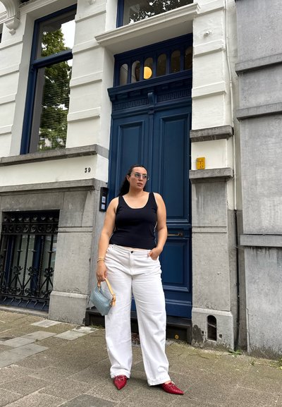 Mujer con camiseta de tirantes negra, pantalones blancos y zapatos rojos de pie sujetando un bolso pequeño frente a una gran puerta azul en una calle de la ciudad.