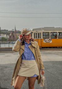 Un trench-coat beige sur une chemise à boutons bleu clair, une jupe crème, une casquette beige et un petit sac à main. Des collants à pois et un arrière-plan urbain.