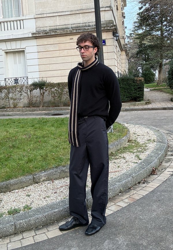 Mann mit lockigem Haar und Brille steht auf dem Bürgersteig neben Gras, trägt schwarzen Pullover, gestreiften Schal, lockere schwarze Hose und schwarze Schuhe.