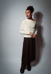 Off-white, asymmetrisch langemouwen top met gedrapeerd detail, gecombineerd met een lange, donkerbruine geplooide rok. Zwarte chunky laarzen. Eenvoudige achtergrond.