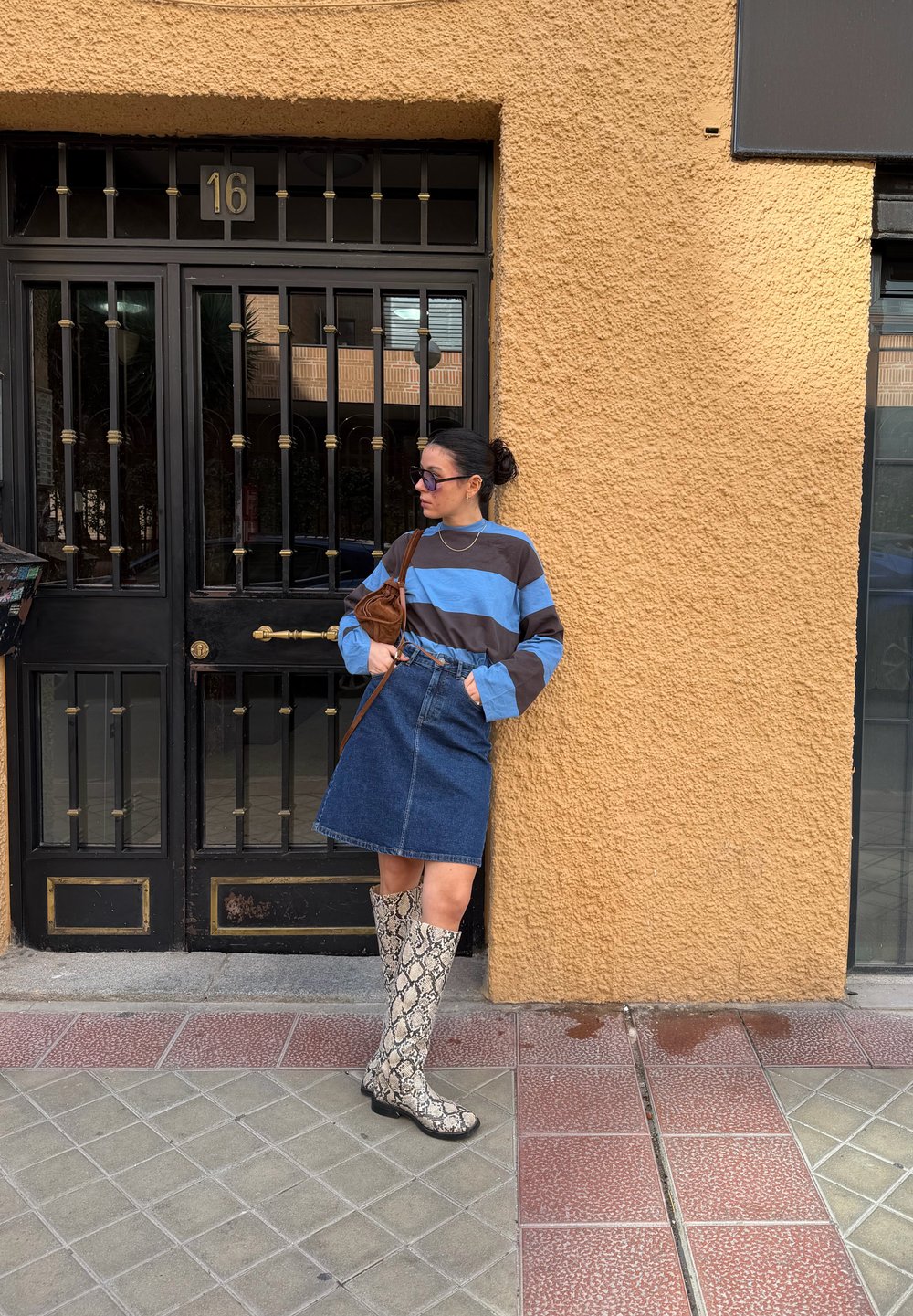 Femme appuyée contre un mur jaune texturé, portant une chemise rayée bleue et marron, une jupe en denim, des bottes montantes à motif serpent, et des lunettes de soleil.