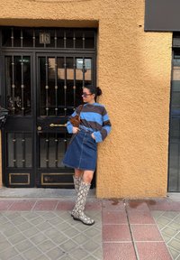 Femme appuyée contre un mur jaune texturé, portant une chemise rayée bleue et marron, une jupe en denim, des bottes montantes à motif serpent, et des lunettes de soleil.