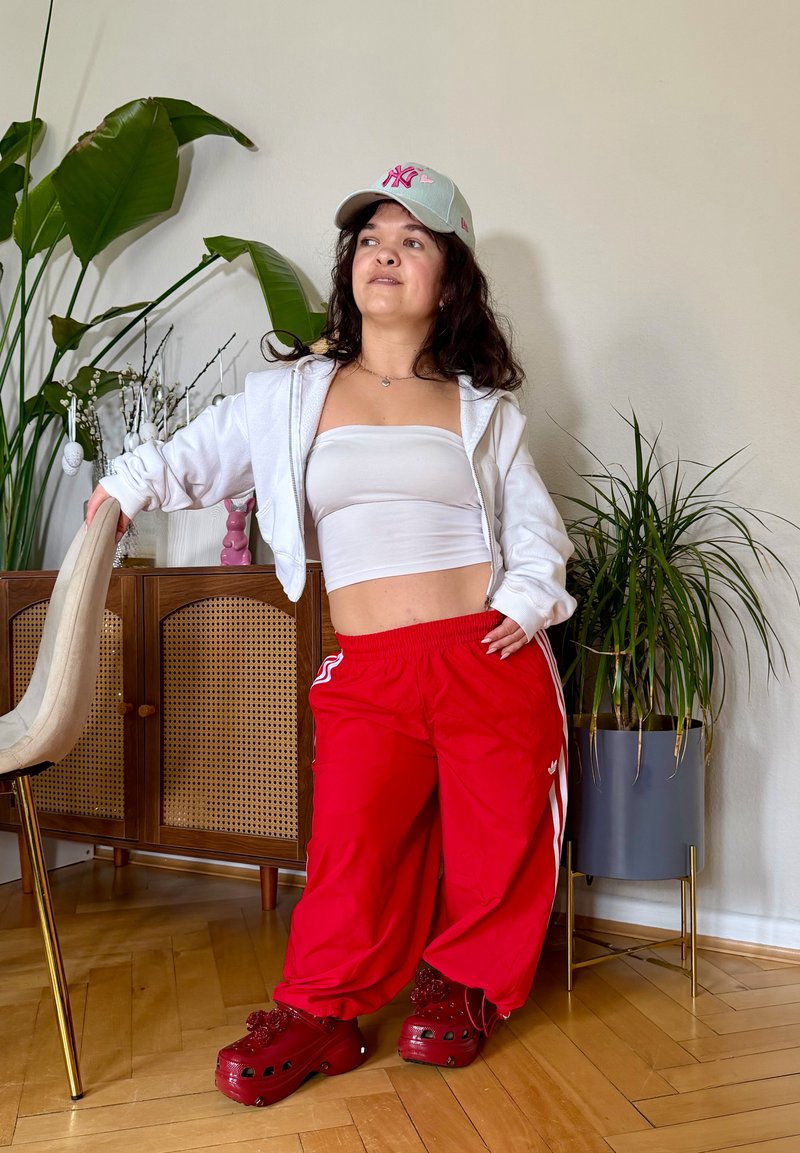 Jeune femme en pantalon rouge, top tube blanc, veste blanche et casquette grise appuyée sur une chaise à côté d'une armoire en bois et de grandes plantes vertes à l'intérieur.