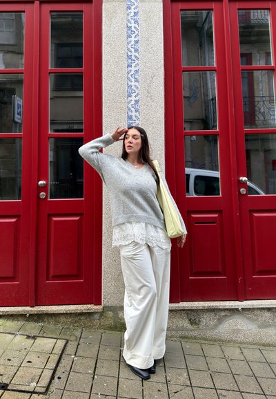 Mujer de pie en la acera frente a puertas con paneles rojos, llevando un suéter gris, blusa blanca de encaje, pantalones anchos blancos y sosteniendo un bolso de mano amarillo.