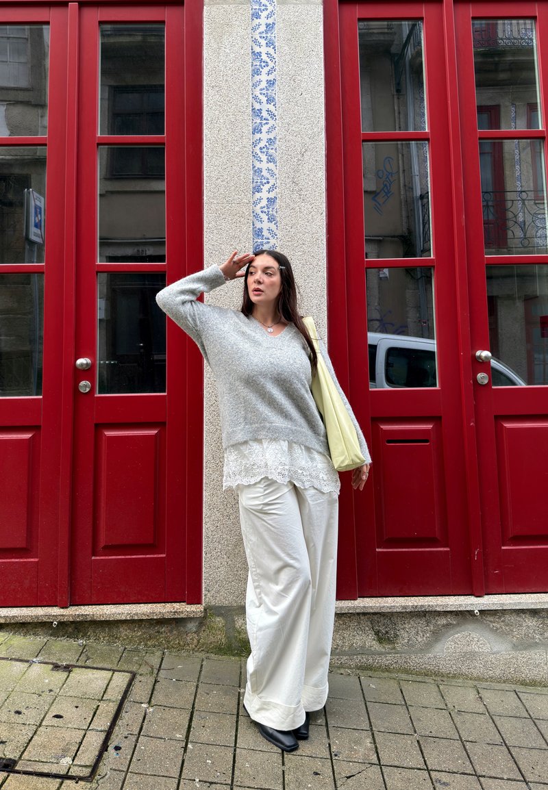 Femme debout sur le trottoir devant des portes à panneaux rouges, portant un pull gris, un chemisier en dentelle blanche, un pantalon large blanc, et tenant un sac fourre-tout jaune.