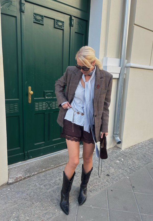 Femme portant des lunettes de soleil, un blazer à carreaux, un haut bleu clair, un short en dentelle noire et des bottes noires, debout sur un trottoir en pierre près d'une porte verte.
