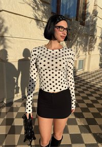 Femme aux cheveux noirs coupés au carré et portant des lunettes, vêtue d'un haut blanc à manches longues à pois, d'une mini-jupe noire, et tenant un sac à main noir en extérieur.