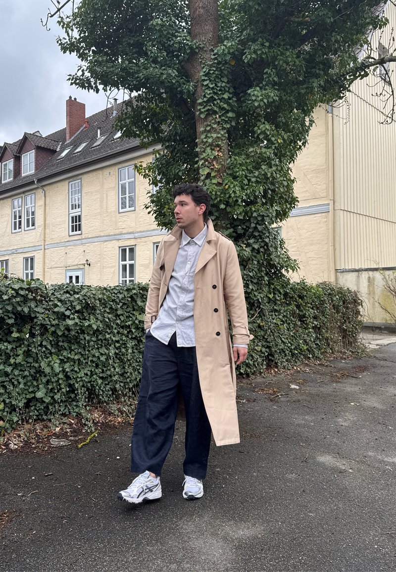Jeune homme portant un trench-coat beige, une chemise blanche et un pantalon bleu marine, debout sur le trottoir près d'un arbre couvert de lierre et d'un bâtiment jaune.