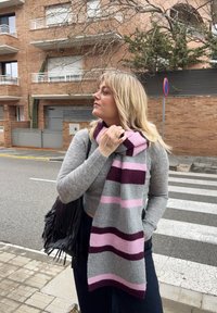 Femme aux cheveux blonds portant un pull gris et une écharpe rayée grise, rose et violette, se tenant près d'un passage pour piétons dans une rue de la ville.
