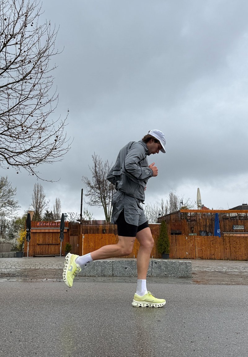 Runner in grey jacket, black shorts, white cap, and yellow shoes jogging on wet pavement with bare trees and wooden fence in background under cloudy sky.