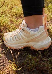 Shoe de caminhada leve com uma palete de cores creme e pêssego, parte superior texturizada, sistema de atacadores seguro e sola robusta para tração.
