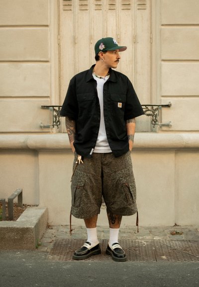 Hombre con tatuajes que lleva una gorra verde, una camisa negra de manga corta, una camiseta blanca, pantalones cortos cargo de camuflaje, calcetines blancos y mocasines negros, de pie junto a una pared de la ciudad.