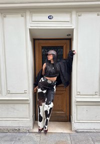 Bukser med sort-hvidt ko-mønster, sort cropped top, oversized sort pufferjakke, brun kasket, sølvspænde, spidse hæle, trædør i baggrunden.