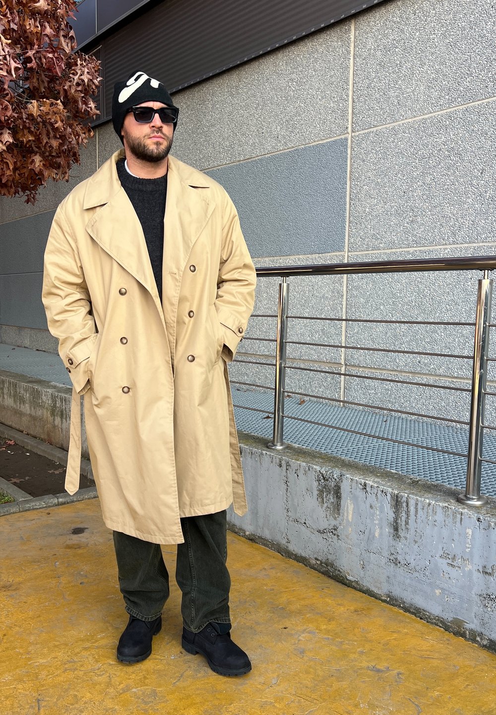 Beige, dupla gombos trench kabát fekete pulóver felett, olívazöld nadrágban és fekete csizmában. Fekete sapka fehér mintával; kültéri környezet.