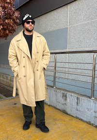 Beige, dupla gombos trench kabát fekete pulóver felett, olívazöld nadrágban és fekete csizmában. Fekete sapka fehér mintával; kültéri környezet.
