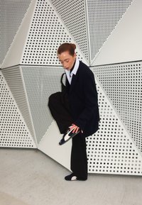 Svarta vida byxor, vit skjorta med krage och en överdimensionerad svart blazer. Skorna har en vit och blå design med en guldaccent.