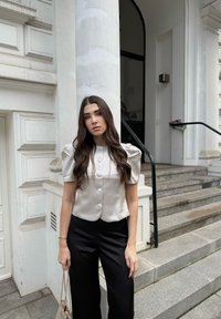 Chemisier boutonné à manches courtes en satin beige clair avec des manches bouffantes, associé à un pantalon large noir. Boutons distinctifs et texture lisse.