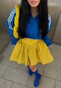 Giacca blu con zip e strisce bianche, gonna plissettata gialla e scarpe blu. Abbinata a una collana di perle e capelli neri lunghi.