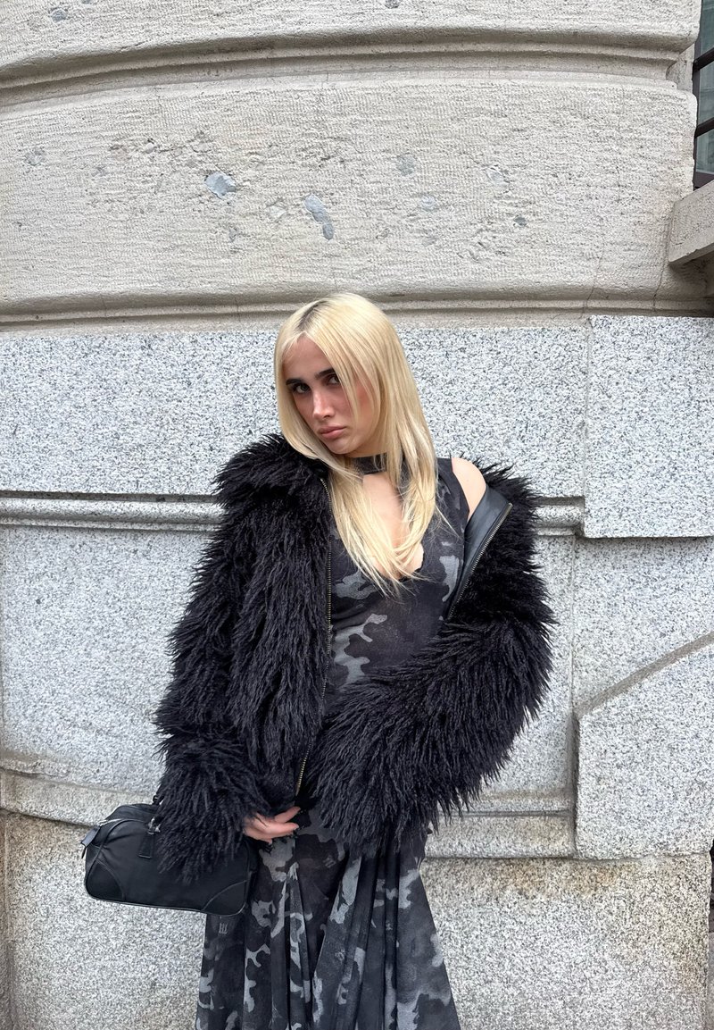 Personne blonde portant une veste noire en fourrure sur une robe camouflage grise, tenant un sac à main noir, debout contre un mur de bâtiment en pierre.