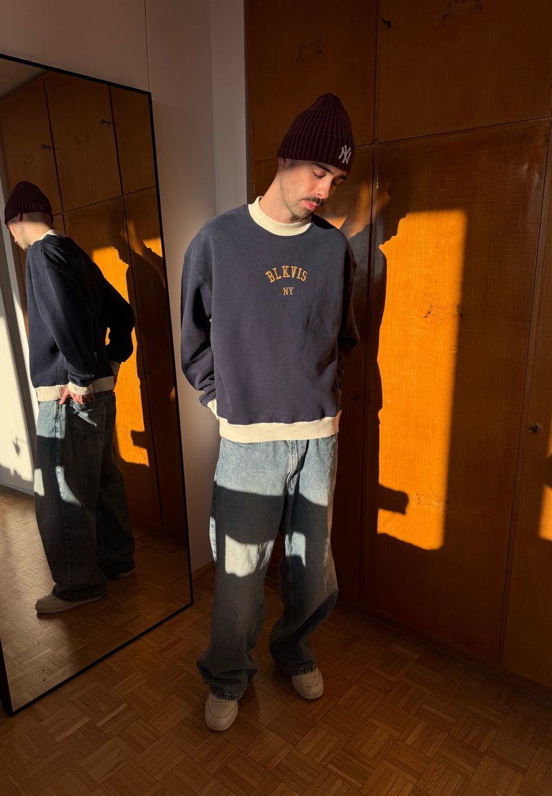 Hombre con gorro de lana, sudadera holgada y vaqueros anchos, de pie con las manos en los bolsillos, junto a un espejo que refleja su espalda.