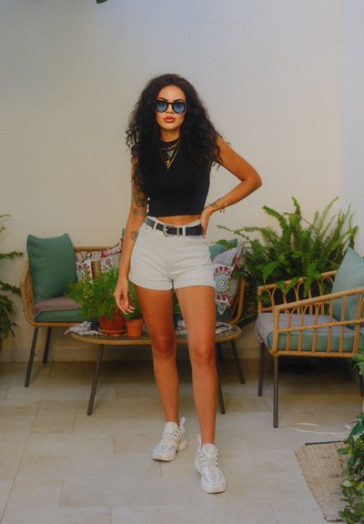 Joven mujer con cabello rizado, usando gafas de sol, top corto negro, pantalones cortos blancos y zapatillas, de pie en el interior cerca de muebles de mimbre y plantas en macetas.